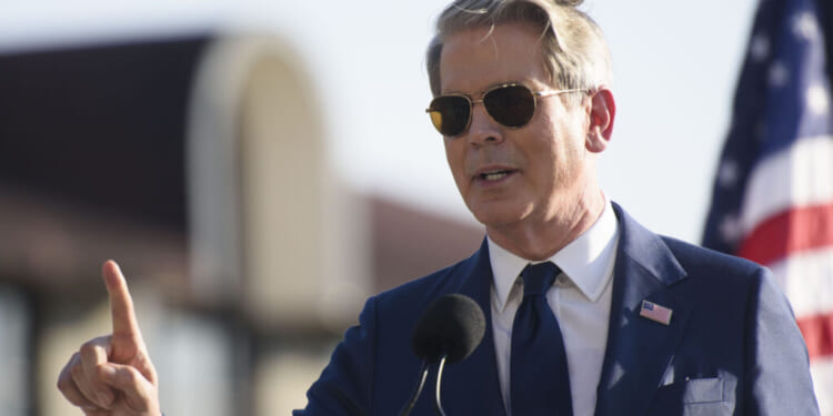 A man in a blue suit and sunglasses speaks at a podium outdoors, gesturing with one finger raised. An American flag is visible in the background.
