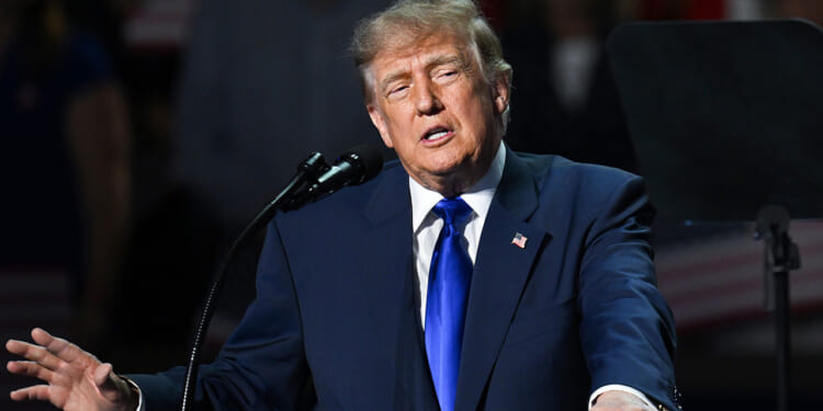 A man in a dark suit and blue tie speaks at a podium with a microphone, gesturing with his hand. An American flag pin is visible on his lapel. The background is dark and blurred.