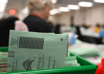 A close-up of green mail-in ballot envelopes in a tray at a busy, blurred election processing facility, with workers in the background.