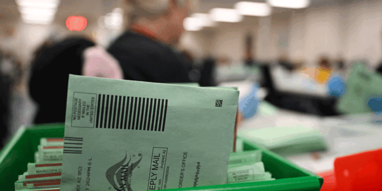 A close-up of green mail-in ballot envelopes in a tray at a busy, blurred election processing facility, with workers in the background.