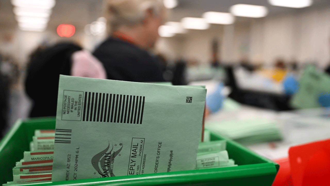 A close-up of green mail-in ballot envelopes in a tray at a busy, blurred election processing facility, with workers in the background.