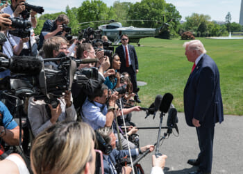 A man in a suit speaks to a crowd of journalists with cameras and microphones on a lawn, with a helicopter and trees in the background.