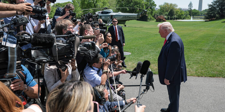 A man in a suit speaks to a crowd of journalists with cameras and microphones on a lawn, with a helicopter and trees in the background.