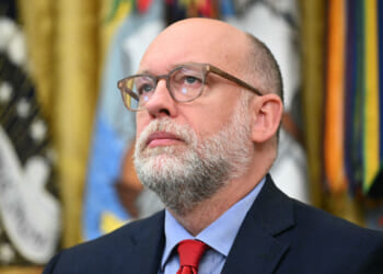Russell Vought with glasses, a gray beard, and a suit with a red tie looks upward. Colorful flags and a blurred emblem are visible in the background. In the Oval Office.