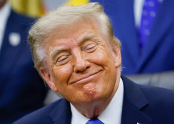Donald Trump in a suit and blue tie sits, smiling with eyes closed, in a brightly lit room. Others in suits are blurred in the background.
