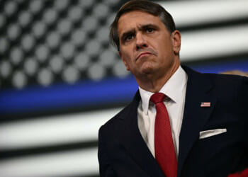 Todd Blanche in a dark suit and red tie stands in front of a blurred black-and-white American flag, looking serious.