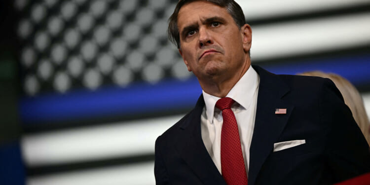 Todd Blanche in a dark suit and red tie stands in front of a blurred black-and-white American flag, looking serious.