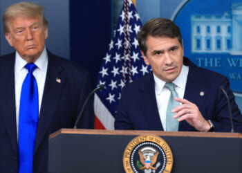 Two men in suits stand at a podium with the U.S. presidential seal. An American flag and a backdrop with the White House logo are visible behind them. One man speaks while the other looks on.
