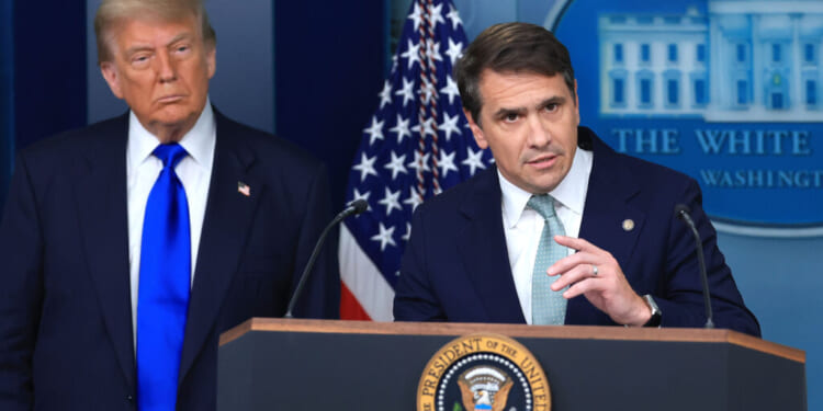 Two men in suits stand at a podium with the U.S. presidential seal. An American flag and a backdrop with the White House logo are visible behind them. One man speaks while the other looks on.