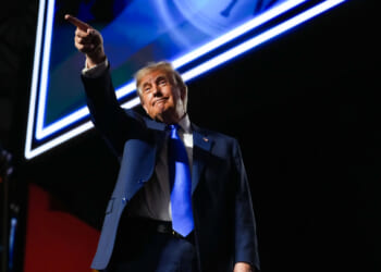 A man in a blue suit and tie stands under bright lights, pointing with his right hand while looking forward. A large screen with a clock image is visible in the background.
