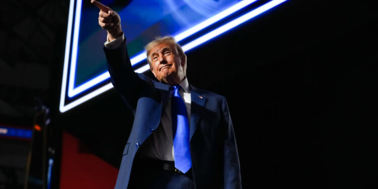 A man in a blue suit and tie stands under bright lights, pointing with his right hand while looking forward. A large screen with a clock image is visible in the background.