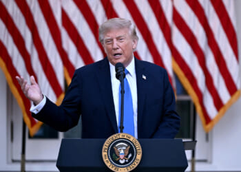 President Donald Trump in a suit speaks at a podium with the U.S. presidential seal, in front of several large American flags.