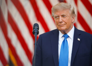 Donald Trump in a dark suit and blue tie stands in front of a microphone, with large red and white striped flags in the background.
