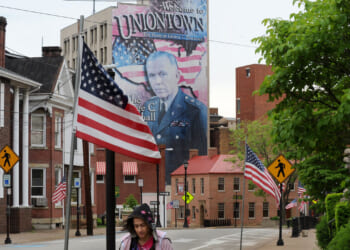A street in Uniontown with American flags, crosswalk signs, and a mural of a man in military uniform with Welcome to Uniontown text in the background. In the distance, part of a power plant rises above the rooftops as a person walks in the foreground.