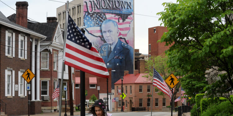 A street in Uniontown with American flags, crosswalk signs, and a mural of a man in military uniform with Welcome to Uniontown text in the background. In the distance, part of a power plant rises above the rooftops as a person walks in the foreground.