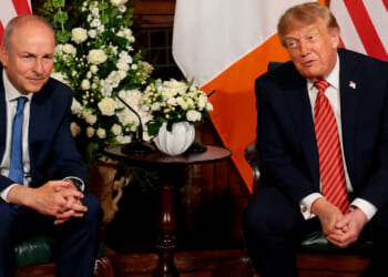 Taoiseach Michael Martin and Donald Trump in suits sit next to each other in front of flower arrangements and the U.S. and Irish flags, engaged in conversation, reflecting the strong diplomatic ties between the United States and Ireland.