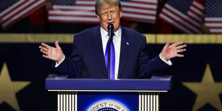 Donald Trump in a blue suit speaks at a podium with the presidential seal, gesturing with both hands, while American flags are held in the background.
