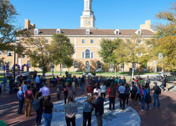 A crowd gathers in a plaza with trees, facing a speaker near balloons. In the background, a large building with a clock tower stands under a clear blue sky.