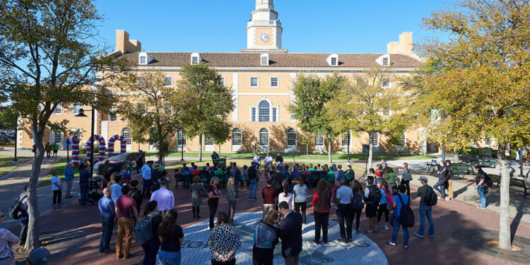 A crowd gathers in a plaza with trees, facing a speaker near balloons. In the background, a large building with a clock tower stands under a clear blue sky.
