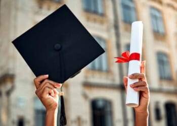 A student raises their degree and their graduation cap at a college campus.