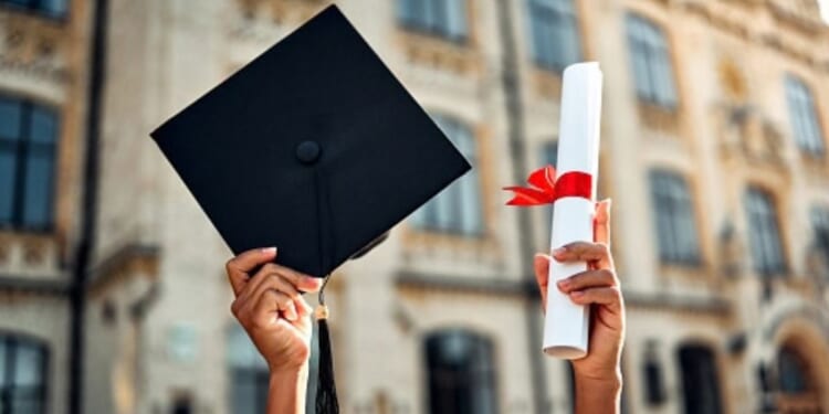 A student raises their degree and their graduation cap at a college campus.