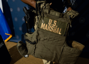 A person holds a tactical vest labeled U.S. Marshal in large yellow letters, with a radio attached. A U.S. Marshals shield and blue flag are visible in the background.