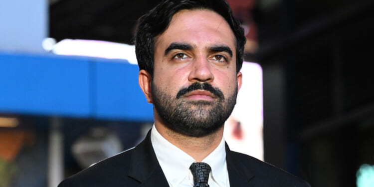 A man with dark hair and a beard wears a black suit, white shirt, and patterned tie, standing outdoors with a blurred city background.