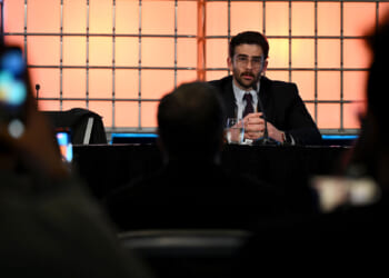Hasan Piker in a suit speaks into a microphone at a table on stage, facing an audience. The background is illuminated with orange grid-like lights, creating a dynamic atmosphere.