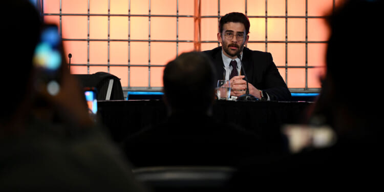 Hasan Piker in a suit speaks into a microphone at a table on stage, facing an audience. The background is illuminated with orange grid-like lights, creating a dynamic atmosphere.