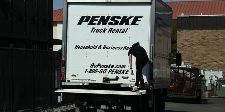 A person climbs onto the back of a white Penske moving truck parked near a building during daylight. The truck advertises household and business rentals, with contact information and a website visible, suggesting it’s ready to support any move—whether for an individual or a union organizing collective effort.