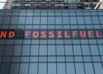 A digital sign on a glass building reads END FOSSIL FUEL$ in bright orange letters, with the S in FUEL$ replaced by a dollar sign.