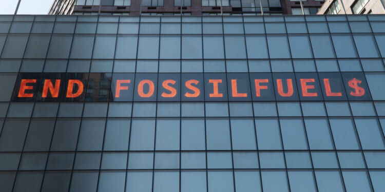 A digital sign on a glass building reads END FOSSIL FUEL$ in bright orange letters, with the S in FUEL$ replaced by a dollar sign.