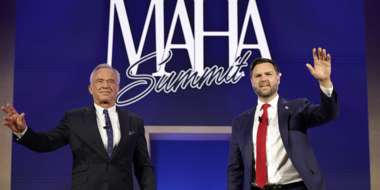 Two men in suits stand on stage, waving, in front of a blue backdrop with MAHA Summit written in large white letters.