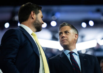 JD Vance and Jim Banks in suits stand close together, facing each other and talking, with a blurred crowd and bright lights in the background.