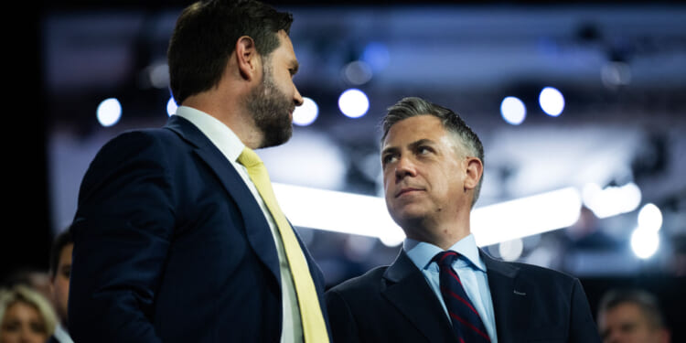 JD Vance and Jim Banks in suits stand close together, facing each other and talking, with a blurred crowd and bright lights in the background.