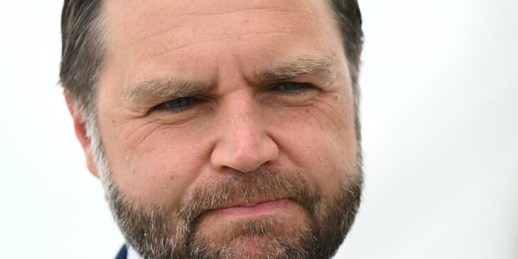 A close-up of JD Vance with short brown hair, a full beard, and a serious expression, with a blurred light background.