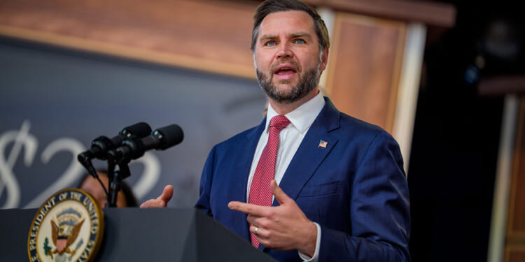 JD Vance in a blue suit and red tie speaks at a podium with microphones; the podium displays the seal of the President of the United States.