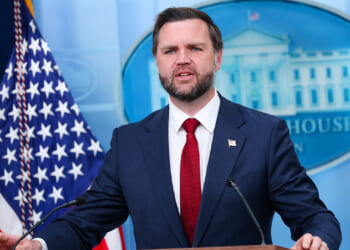 JD Vance in a dark suit and red tie speaks at a podium in front of American flags and a White House emblem backdrop.