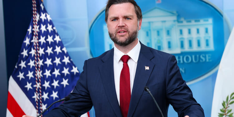 JD Vance in a dark suit and red tie speaks at a podium in front of American flags and a White House emblem backdrop.