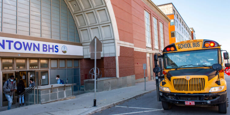 A yellow school bus is parked outside a brick school building labeled DOWNTOWN BHS, while students enter through the main doors. The weather is clear and sunny.