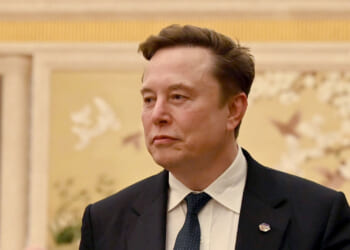 A man in a dark suit and blue tie stands indoors with a neutral expression. The background features a light-colored wall with floral and bird artwork.