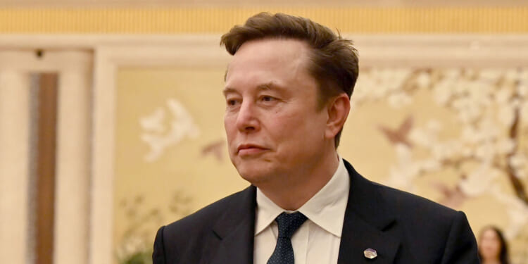 A man in a dark suit and blue tie stands indoors with a neutral expression. The background features a light-colored wall with floral and bird artwork.