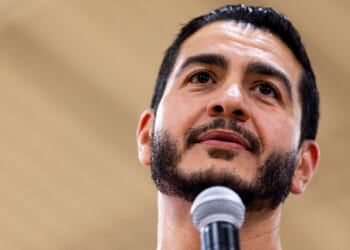 A man with short dark hair and a beard speaks into a microphone, looking slightly upward, with a blurred beige background.