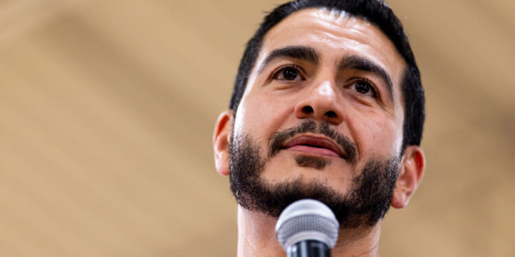 A man with short dark hair and a beard speaks into a microphone, looking slightly upward, with a blurred beige background.