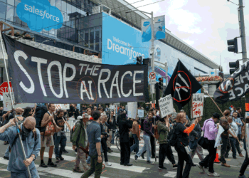 Protesters cross a city street holding signs, including a large banner reading Stop the AI Race, in front of a modern building with Salesforce and Dreamforce banners.