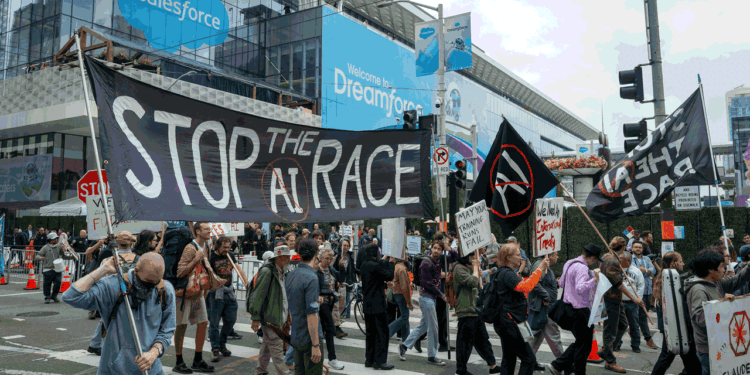 Protesters cross a city street holding signs, including a large banner reading Stop the AI Race, in front of a modern building with Salesforce and Dreamforce banners.