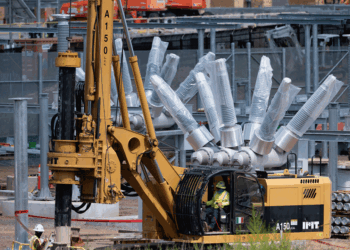 A worker operates a yellow drilling machine at a construction site, with large metallic components and equipment visible in the background.