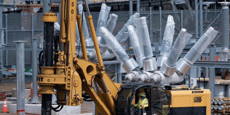 A worker operates a yellow drilling machine at a construction site, with large metallic components and equipment visible in the background.