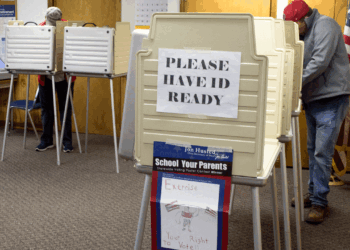 Two people vote at booth stations in a polling place, with a large sign reading PLEASE HAVE ID READY displayed. An American flag stands in the corner. The scene suggests a formal voting environment.