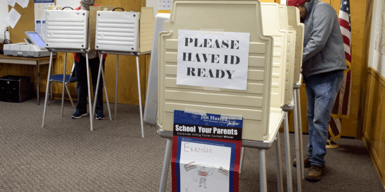 Two people vote at booth stations in a polling place, with a large sign reading PLEASE HAVE ID READY displayed. An American flag stands in the corner. The scene suggests a formal voting environment.